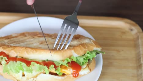 A sandwich on a plate being sliced with a fork and knife on a wooden board.