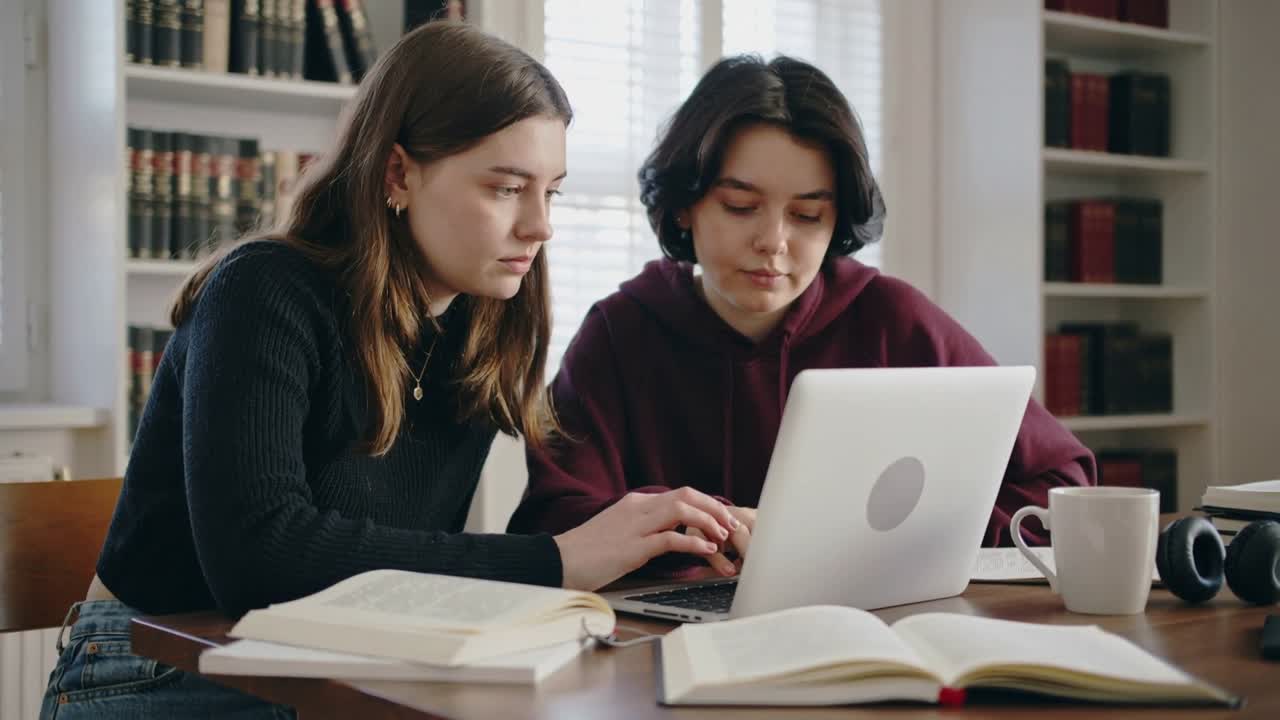 Legger hendene på laptop, personen til venstre iført svart genser taster ved bordet, mens studenten til høyre ser på bøker.