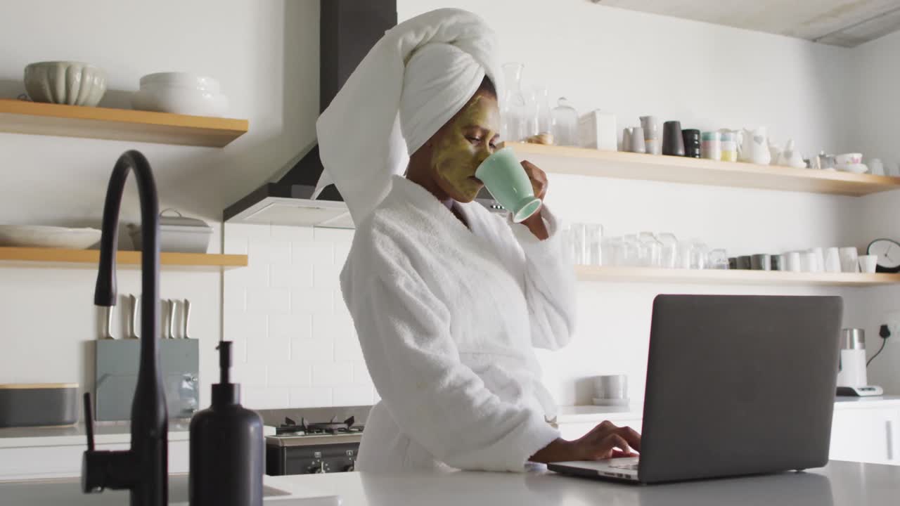 feliz mujer afroamericana con máscara de belleza en la cara, bebiendo café y usando computadora portátil en la cocina