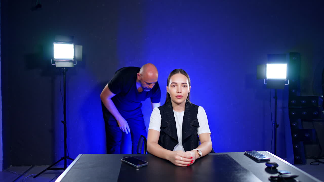 Focused Caucasian woman with long brunette hair sits at the desk in studio. Male photographer sets light at backdrop.
