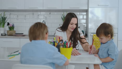 Mom-helps-sons-learn-to-draw-doing-homework-preschool-preparation-at-home-sitting-in-the-white-kitchen.-Two-brothers-draw-a-portrait-of-mom-together