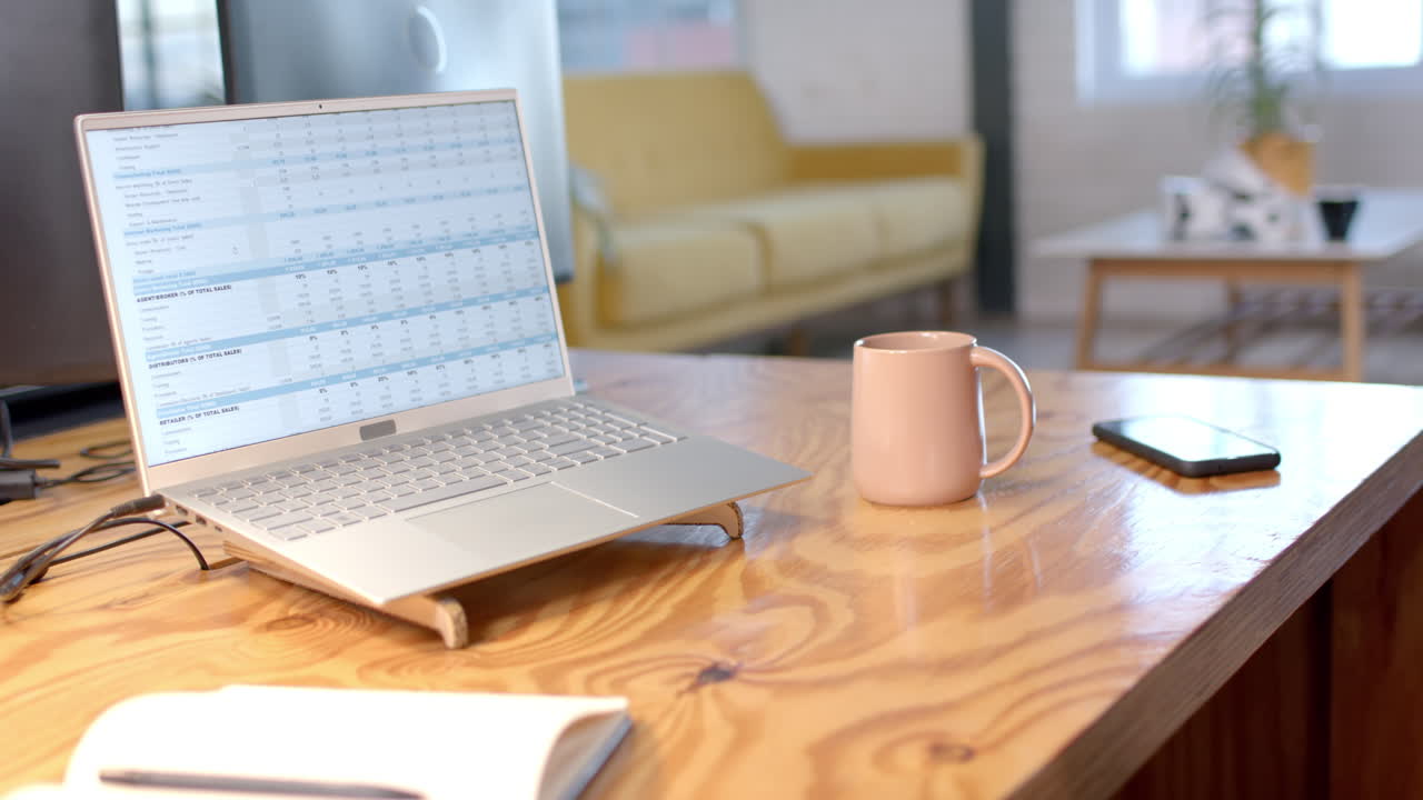 Laptop displaying sales tracker on wooden table, pink mug and smartphone beside