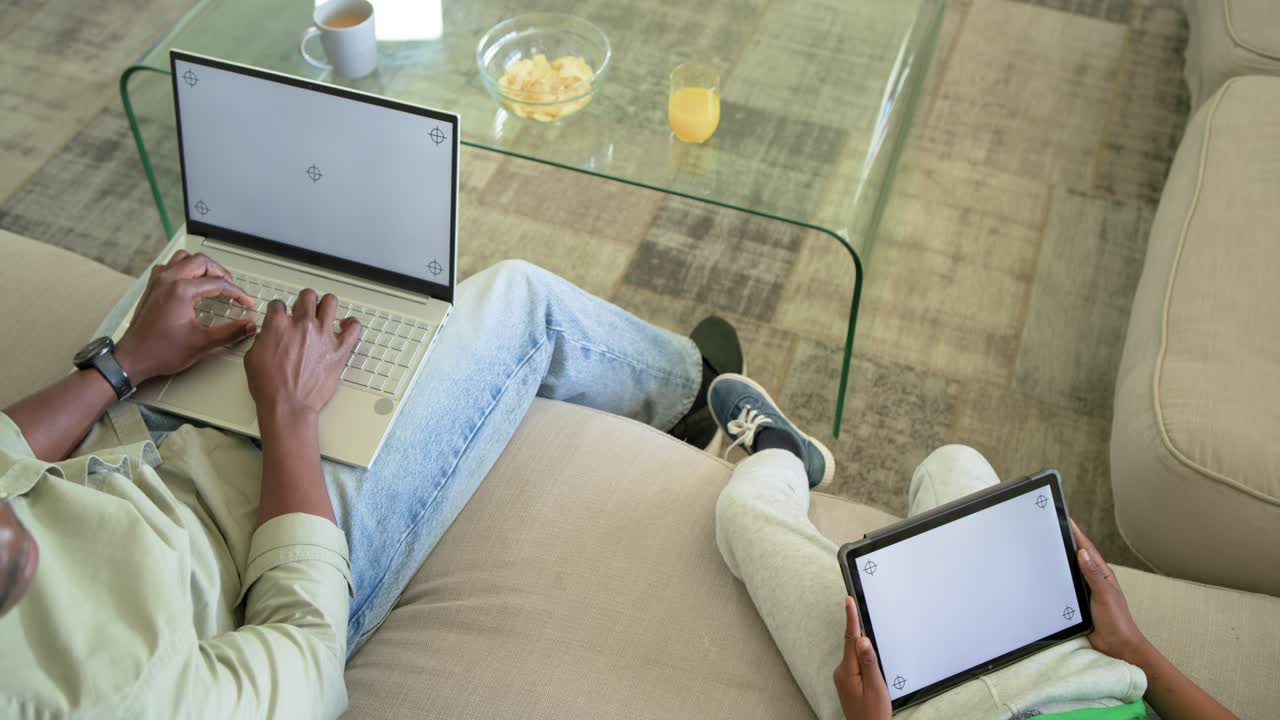 African American father and son typing on laptop while testing tablet screens on couch with snacks