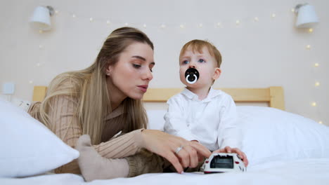 beautiful caucasian mother lies on the bed near her little son. baby boy plays with a toy car.