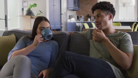 Happy-biracial-couple-sitting-on-sofa-in-living-room-drinking-coffee