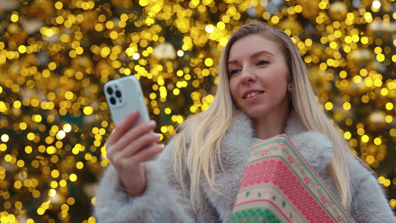 Woman with gifts taking selfie in front of Christmas lights