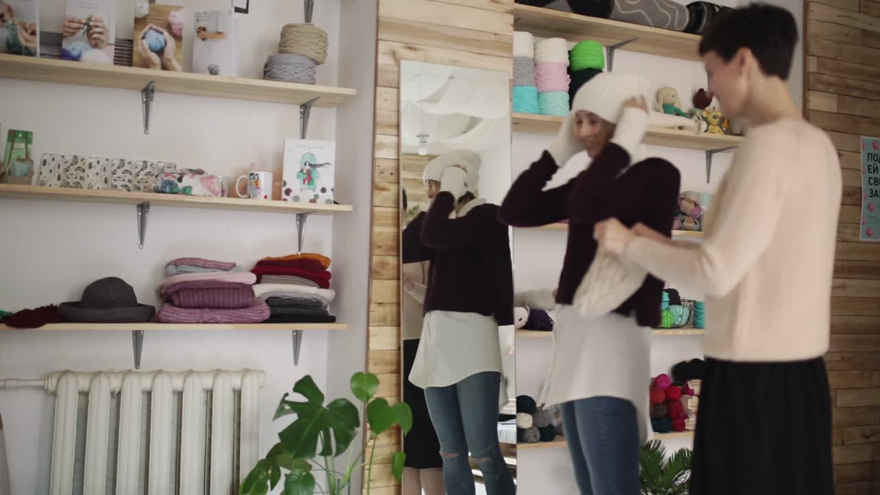 젊은 여자는 작은 가게 앞 거울에 니트 모자를 끼고 있다. 작업장에 있는 여자