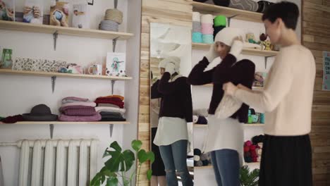 Una-Mujer-Joven-Se-Coloca-Un-Gorro-De-Punto-Frente-Al-Espejo-En-Una-Pequeña-Tienda.-Mujer-En-Taller