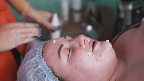 beautician-applies-moisturizer-onto-large-woman-face-closeup