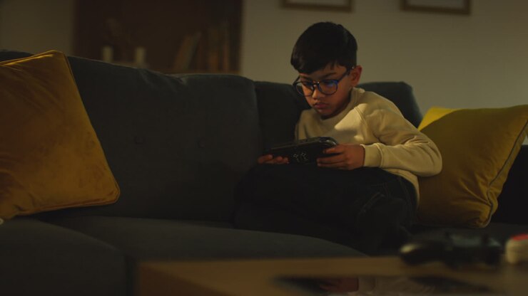 Ragazzo giovane seduto sul divano a casa che gioca o guarda contenuti in streaming su un dispositivo di gioco portatile 7