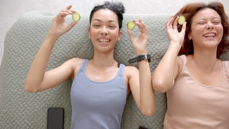 two female friends are lying down with slices of cucumber on their eyes, smiling joyfully