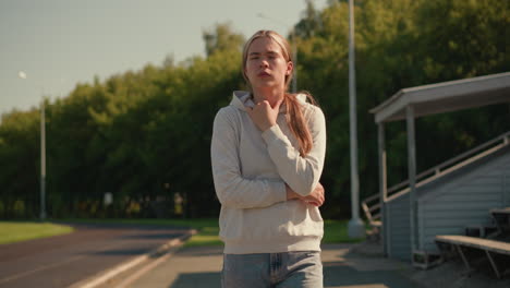 young woman with arms folded, thoughtfully touching her hoodie, walking outdoors near stadium, background includes trees, electric poles, and stadium seating