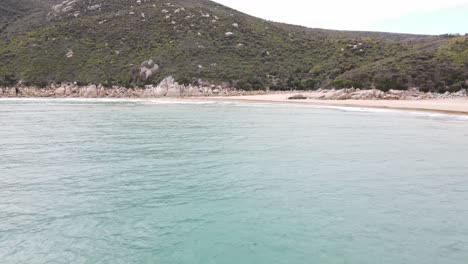 Drone-aerial-hovering-over-beautiful-beach-shoreline-with-blue-water-and-green-mountains-in-Wilsons-Promontory