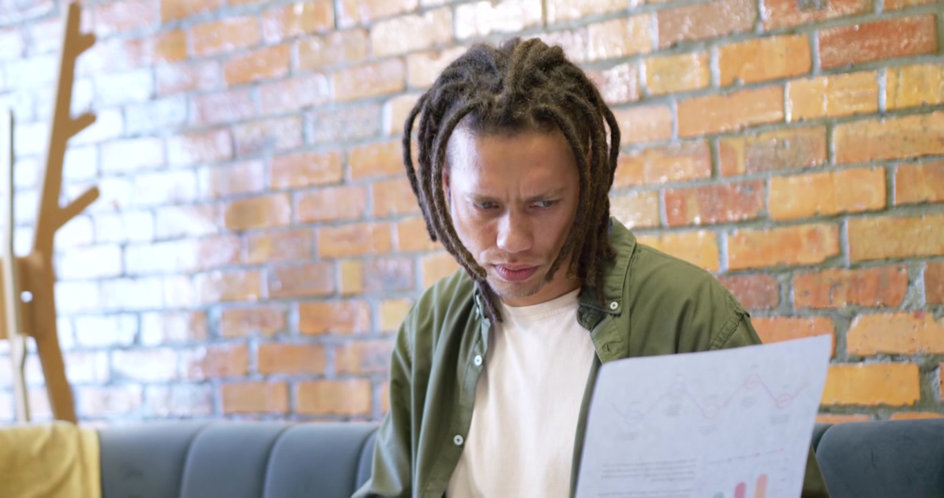Afroamerikansk mand i olivenfarvet skjorte på café læser rapportdiagrammer og verificerer tal på bærbar.
