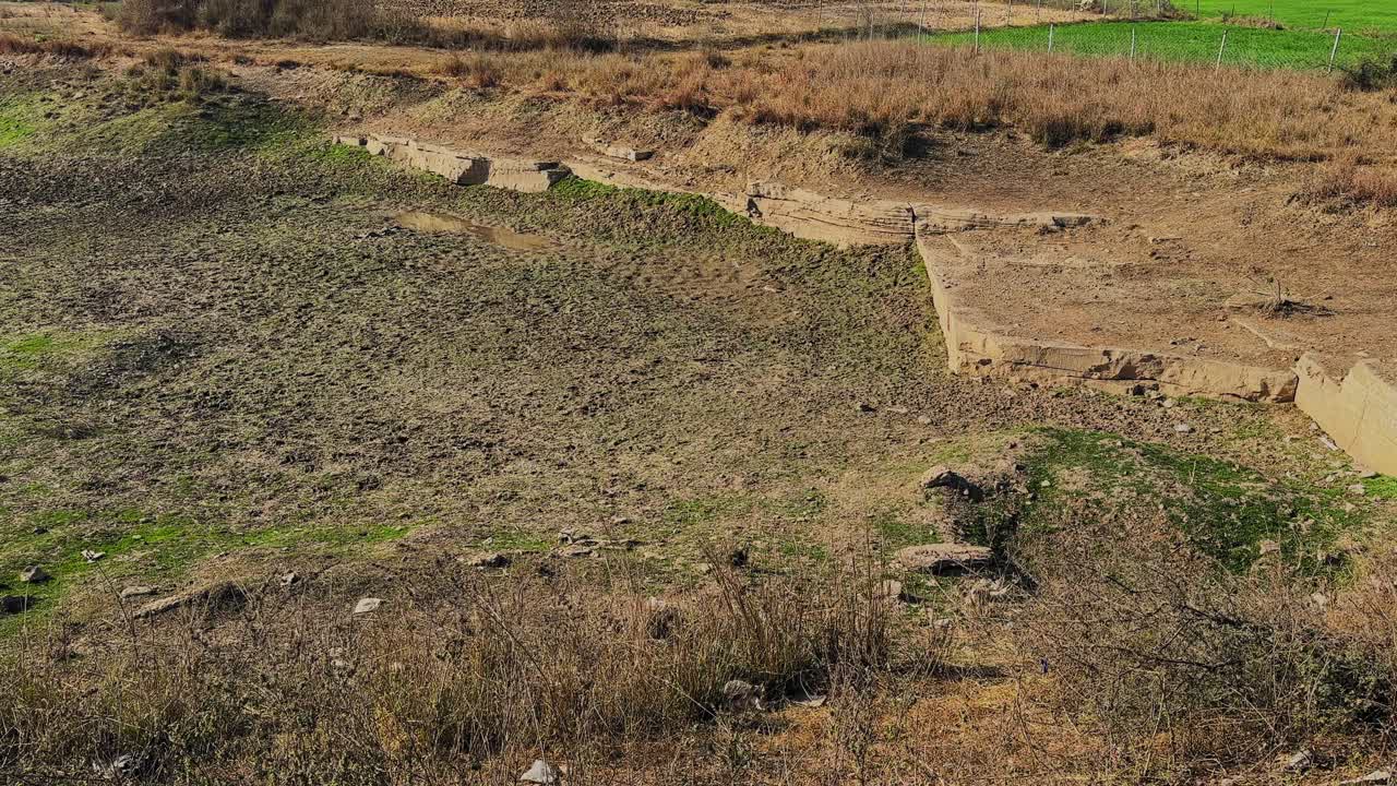 Plan large d’un lit d’étang asséché montrant le sol exposé et les couches de pierre, les textures de sol irrégulières, la faible végétation verte et les bords érodés dans un paysage rural