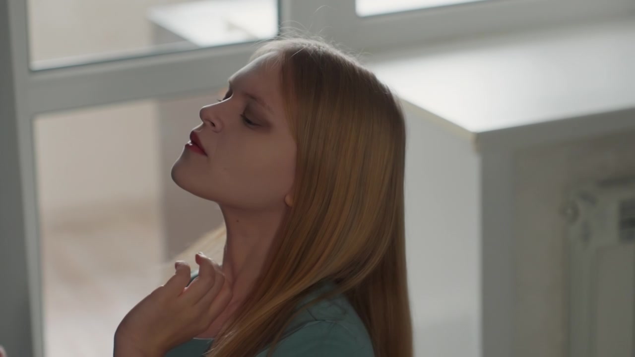 Caucasian woman near window in house tilting head and massaging neck in a quiet selfcare moment. Soft daylight outlines profile and radiator detail, gentle movements show posture adjustment, breathing