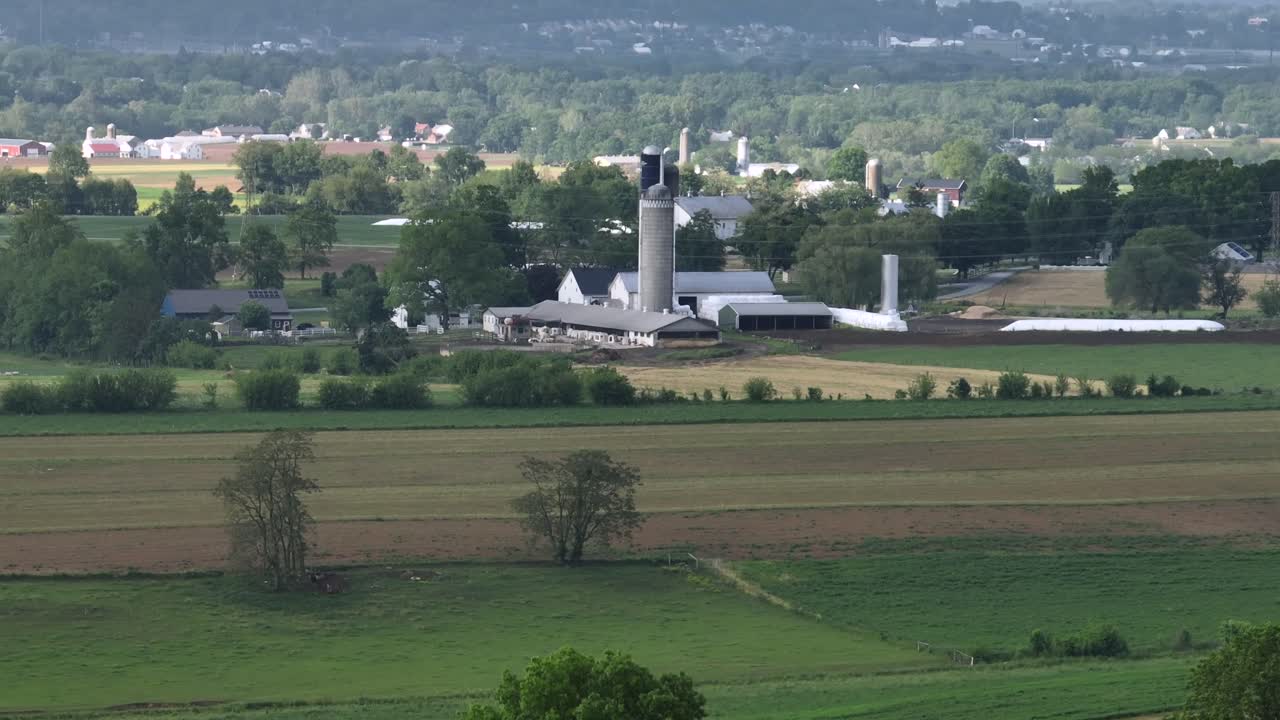 Aerial zoom shot of American farmstead with silo storage in green landscape of United States. Forst landscape and agricultural farm fields at cloudy day.