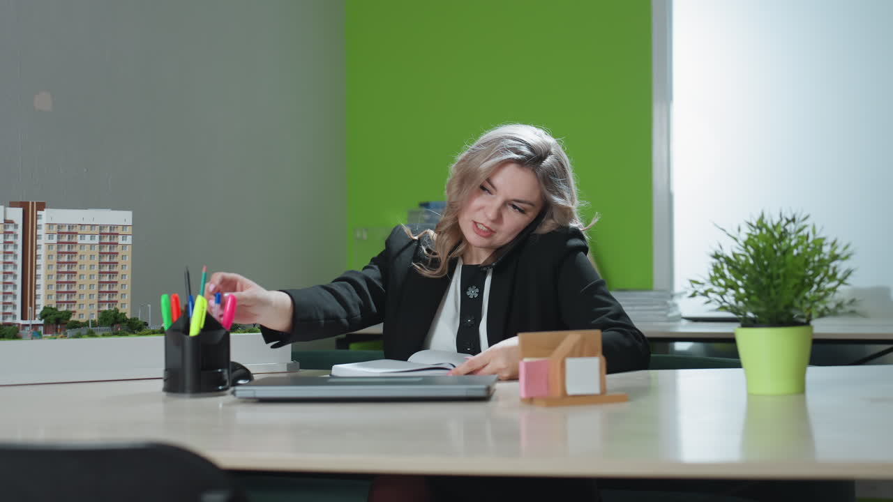 sales agent picks call from friend smiling friendly while opening book and grabbing pen to take notes, multitasking at desk with architectural model and office supplies in modern workspace