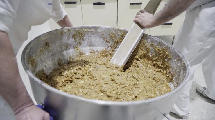 Slow motion handheld shot of a large mixing pot with a mass or dough for candy being stirred by a baker while his assistant holds the pot in a candy factory in medina sidonia