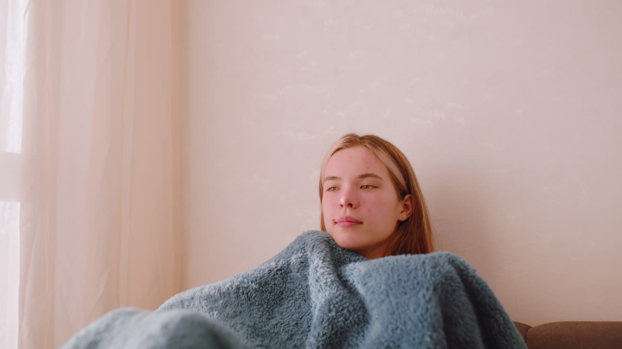 Lady seated on sofa covering herself with blanket while facing window with eyes closed enjoying soft daylight and peaceful comfort of solitude relaxation warmth