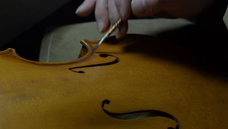skilled violinmaker works on creating an aged appearance around the purfling and f-holes of a violin top plate in workshop setting, captured in close-up detail shot using brush and finger process