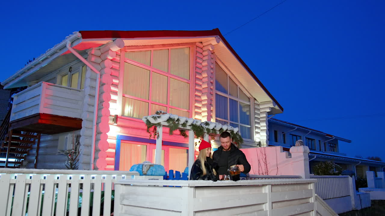 Caucasian couple stands on the porch near a beautiful cottage at sunset. Man pours some tea into cup. Romantic winter evening for two.