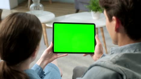 más de la vista del hombro pareja joven en casa sentada en un sofá usando con pantalla de maqueta verde tableta. hombre y mujer usando dispositivo de pantalla táctil, navegación por internet. concepto de pantalla verde y clave de croma