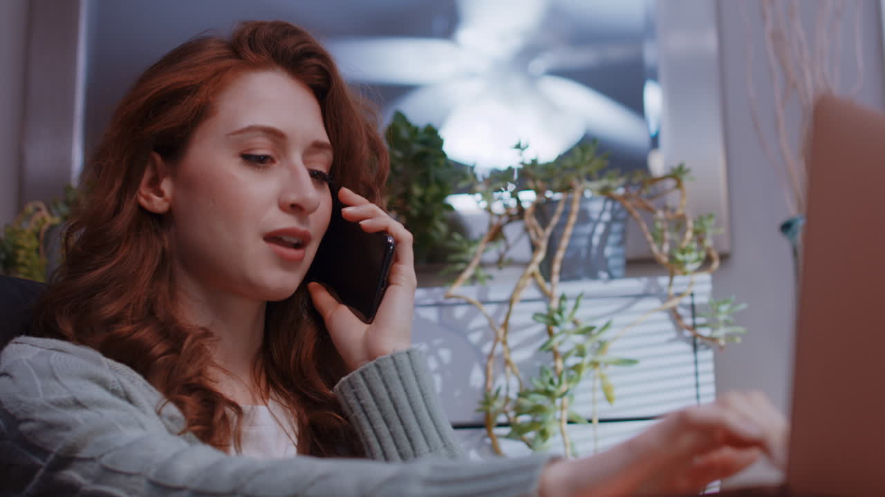 una joven mujer caucásica está trabajando desde casa en su computadora portátil, hablando en su teléfono celular