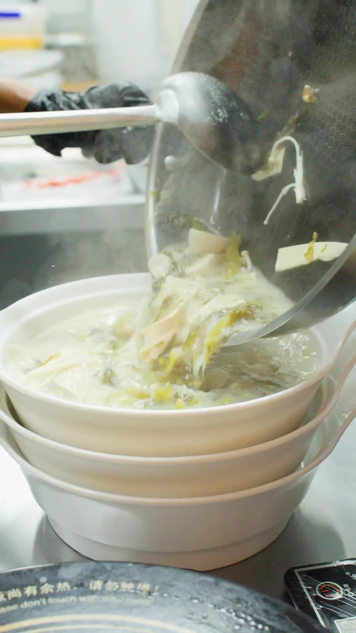 業務用の厨房で、黒い手袋をはめたプロのシェフが、湯気の立つ麺スープを丼に注いでいる。