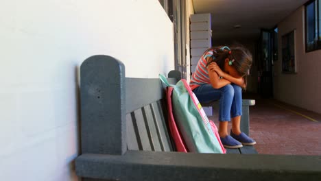 side view of african american schoolgirl with head down sitting alone on bench in school corridor 4k