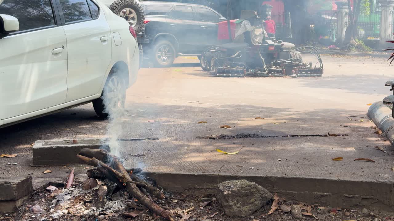 Un piccolo fuoco di legna brucia a terra vicino ad auto parcheggiate in pieno giorno.