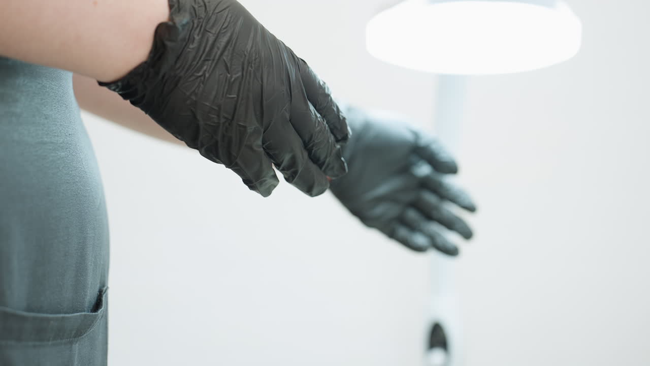 Close up view of beautician gloved hand as she gestures while standing in salon wearing black apron, with background softly blurred and lamp visible, indicating professional client service environment