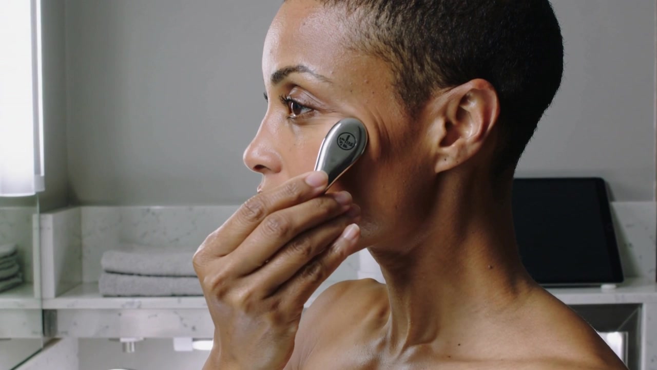 Holding metal facial device, bare-shouldered woman gliding tool along cheek at sink for skincare