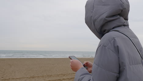 Frau-In-Jacke-Surft-An-Einem-Trüben,-Kalten-Tag-Auf-Dem-Handy-Im-Internet-In-Der-Nähe-Des-Meeres