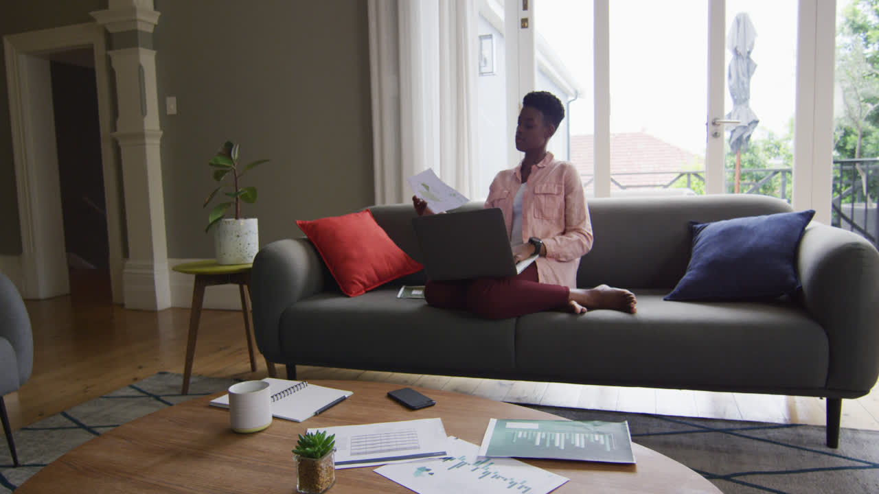 Femme afro-américaine lisant un document et utilisant un ordinateur portable en télétravail