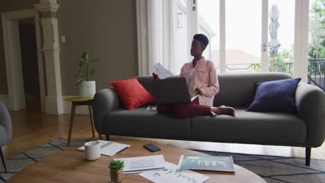 mujer afroamericana leyendo un documento y usando una computadora portátil mientras trabaja desde casa