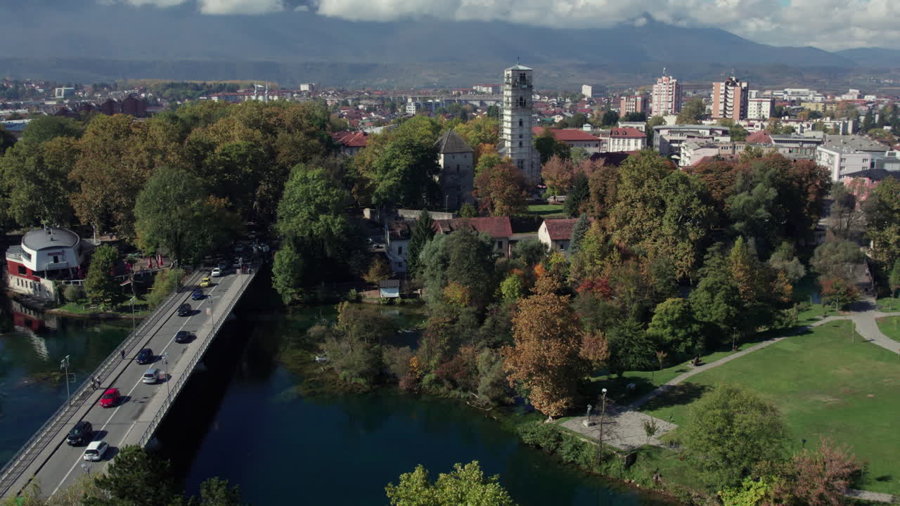 Aerial drone view of Bihać city center in Bosnia and Herzegovina featuring the main bridge over the Una River, colorful autumn trees, historic buildings, and distant mountain scenery under clear skies