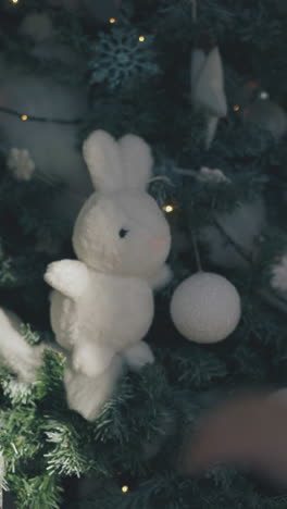 blond long haired little girl puts white stuffed rabbit on decorated christmas tree branch in room close backside view