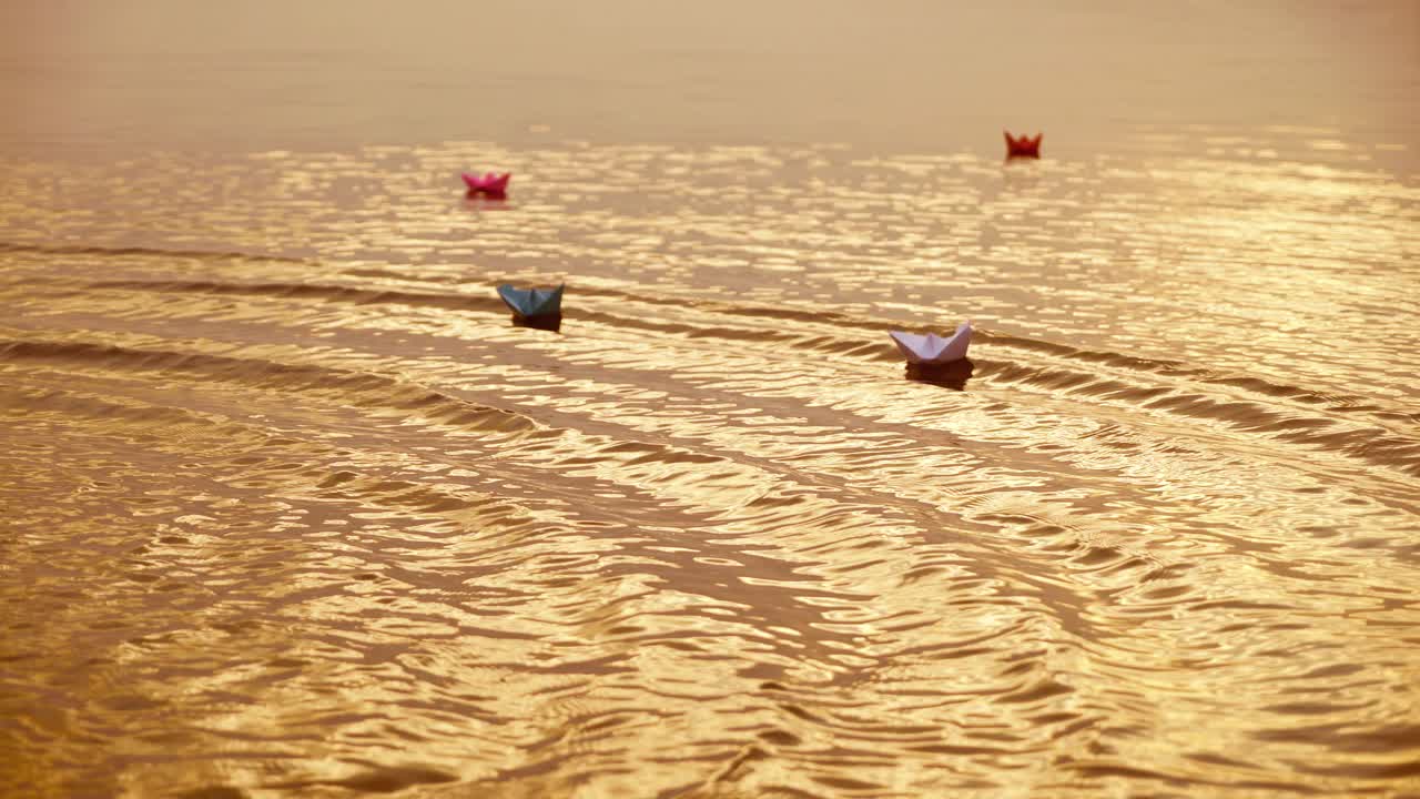 Multy-colored paper boats floating on waves in a river at the evening sunset. Small ships made from paper sail in water.
