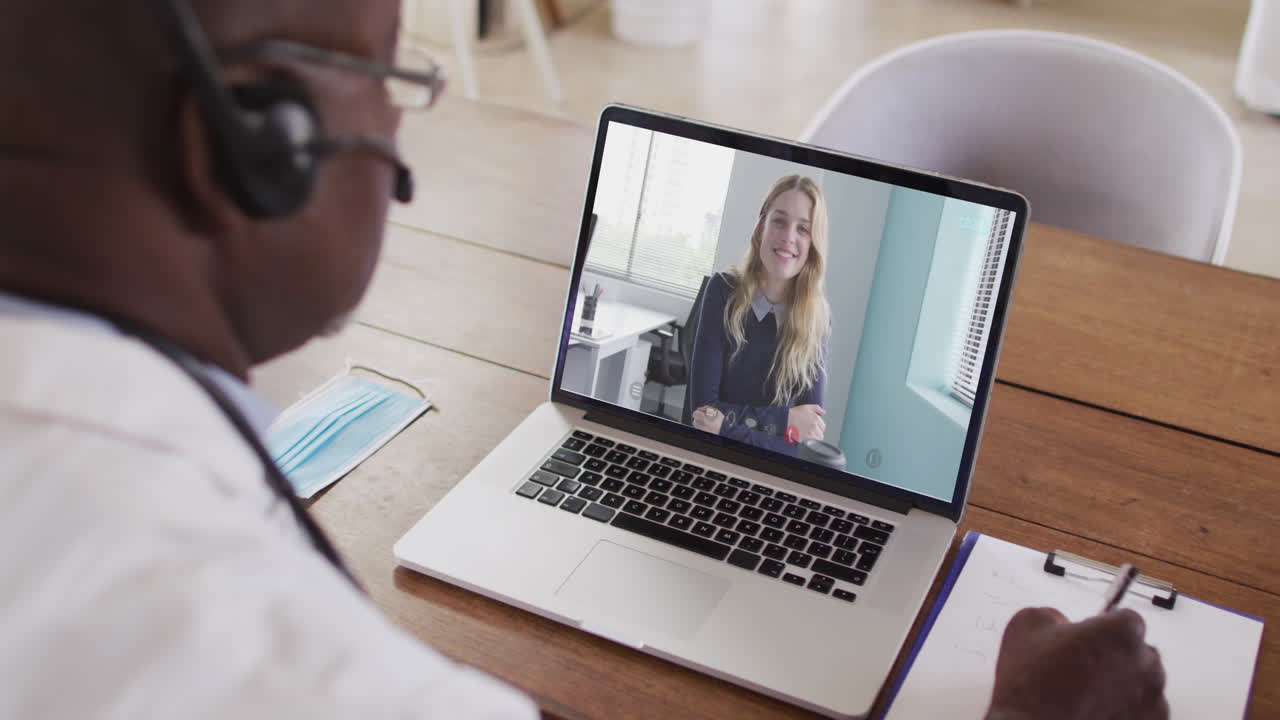 Ein afroamerikanischer Arzt mit Telefon-Headset macht sich Notizen während eines Videoanrufs am Laptop.