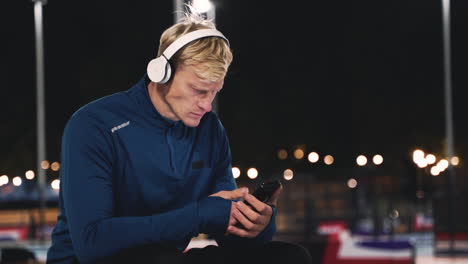 hombre rubio deportivo sentado en el parque escuchando música con auriculares bluetooth y usando el teléfono móvil mientras toma un descanso durante su sesión de entrenamiento por la noche