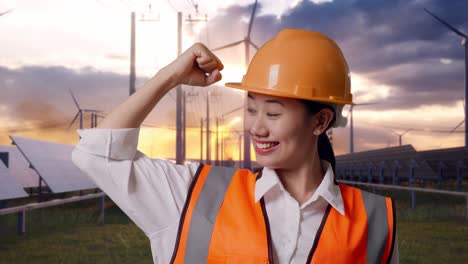 close up of asian female engineer with safety helmet flexing her bicep and smiling to camera with solar panel and wind turbines