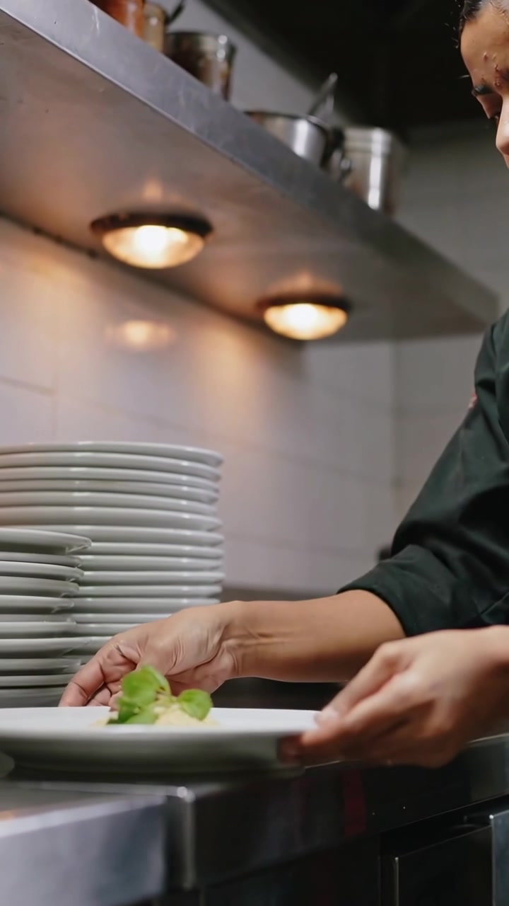 Vertical video: Placing green sprig as plate arrives chef in coat nudging sliding at pass to serve