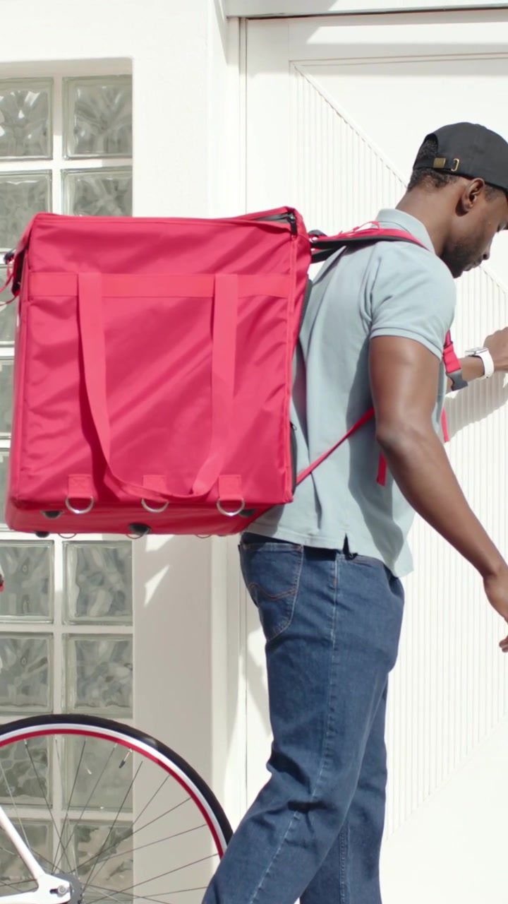 Vertical video: Arriving courier in cap shifting red bag, reaching home delivering meal with bike