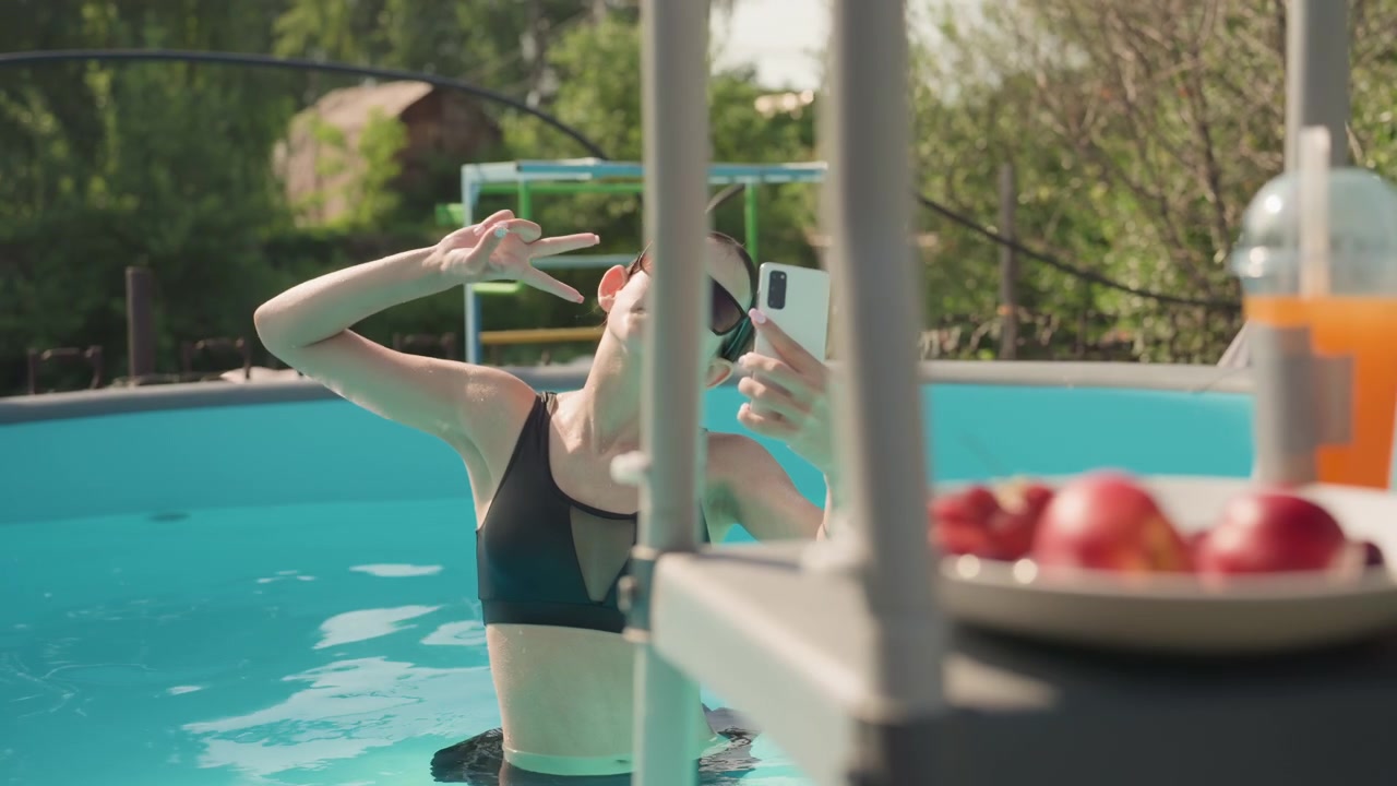 Woman in black sports bra takes selfie with white smartphone while standing waist deep in outdoor swimming pool. Poolside setting includes green foliage metal ladder structure and bright