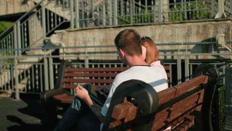 estudiante estudiando al aire libre sentado en un banco como señora muestra tipo algo en la tableta, participando en la discusión, el fondo cuenta con barandilla de hierro, escalera, y la cálida luz del sol iluminando el entorno