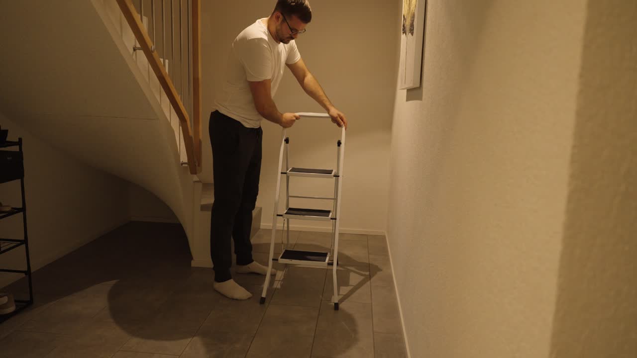 A man unfolds and positions a step ladder, carefully checking its stability and ensuring it’s safe to use in a home hallway