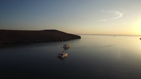 Toma-Aérea-De-Un-Velero-Catamarán-Y-Un-Yate-En-Una-Tranquila-Bahía-Al-Atardecer,-Mar-De-Cortez,-Baja-California-Sur