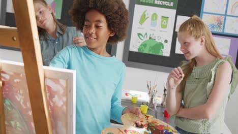 Video-of-diverse,-happy-schoolchildren-standing-at-easels-and-painting-in-art-class,-copy-space