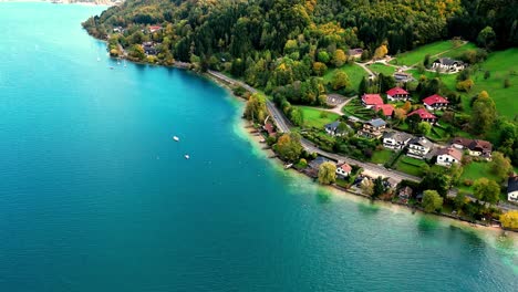 a vibrant summer scene of a long, narrow lake surrounded by lush green mountains, dotted with colorful houses with red roofs and sailboats with white sails, captured from a bird’s-eye view